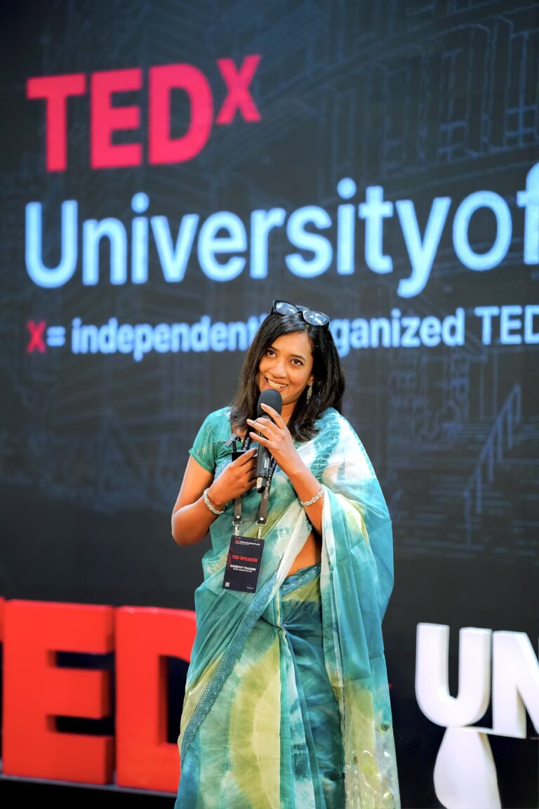 Shurovy Yeasmin delivering her talk at TEDx University of Asia Pacific, wearing a teal saree and holding a microphone.